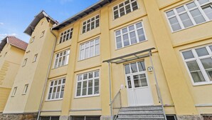 Um edifício de vários andares com fachada amarela, janelas brancas e escadaria de metal que dá acesso à entrada.