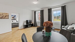 Sala de estar moderna com sofá, televisão, mesa de jantar e vista para uma paisagem verde através de grandes janelas.