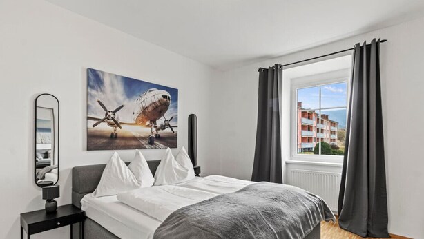 Quarto com cama, janela com cortinas, quadro de um avião e mesa de cabeceira com abajur.