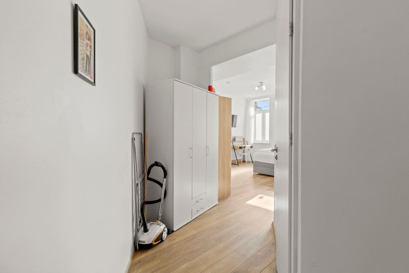 A hallway with a white wardrobe, a vacuum cleaner, and a dining area with a table and chairs.