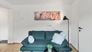 A room with a teal sofa, a white fluffy pillow, a small plant, and a wall-mounted picture of flowers. - FarawayHomes Studio Leoben #P82 (Donawitz)
