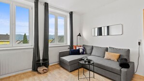 Ein modernes Wohnzimmer mit einem grauen Sofa, einem runden Couchtisch, einer Stehlampe und Blick durch große Fenster ins Freie.