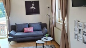 Een woonkamer met een grijze bank, een klein rond tafeltje, een grijze vloerkleed, een foto in een zwarte lijst aan de muur, en een aan de muur bevestigde televisie.
