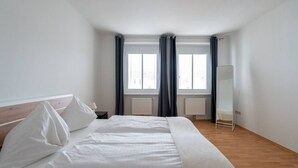 Une chambre à coucher avec un lit, une table de chevet, un miroir et deux fenêtres avec des rideaux.