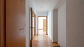 Un couloir au plancher en bois, une porte sur la gauche et, au bout, une porte ouverte qui donne sur une autre pièce.