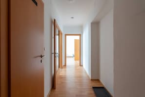 A hallway with wooden flooring, a door on the left, and a view into another room through an open door at the end.
