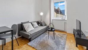 Een moderne woonkamer met een grijze bank, een klein tafeltje met een plant, een televisie op een meubelstuk en een raam met uitzicht op gebouwen en bergen.