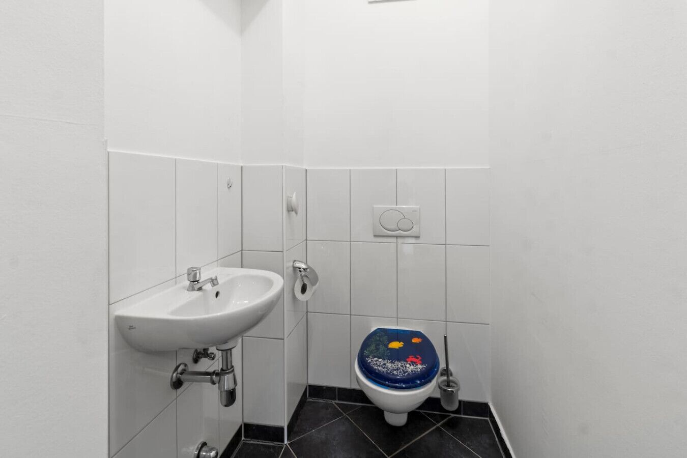 Ein Badezimmer mit einem weißen Waschbecken, einer Toilette mit blauem Deckel und einem an der Wand montierten Spülhebel.