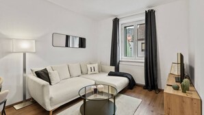 A modern living room with a sofa, a glass coffee table, a lamp, a window with curtains, and a cactus on a wooden side table. - FarawayHomes Studio Fohnsdorf #4 (Fohnsdorf)