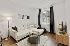 Ein modernes Wohnzimmer mit einer Couch, einem Glastisch, einer Lampe, einem Fenster mit Vorhängen und einem Kaktus auf einem Holztischchen.