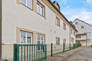 Ein mehrstöckiges Gebäude mit einer beige farbenen Fassade, zahlreichen Fenstern und einem grünen Metallzaun im Vordergrund.