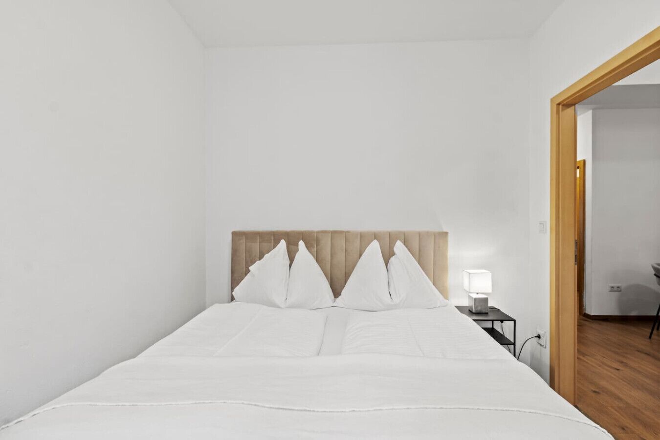 A minimalist bedroom with a bed, white bedding, a wooden headboard, a bedside table with a lamp, and a wooden floor.