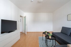 Ein modernes Wohnzimmer mit Fernseher, grauem Sofa, rundem Tisch mit Pflanze und einem gemusterten Teppich.