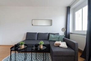 Una sala de estar moderna con un sofá gris, dos mesas auxiliares con plantas en macetas, una lámpara de pie y un ventanal con cortinas.