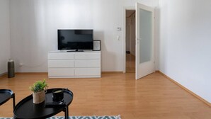 A living room with a TV on a white cabinet, a small table with a plant and a cup, and a door leading to another room.