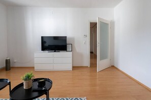 Ein minimalistisches Wohnzimmer mit einem Flachbildfernseher auf einer weißen Kommode, einem kleinen Tisch mit Pflanze und Tasse sowie einer Tür, die in einen anderen Raum führt.