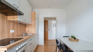 A compact kitchen with wooden cabinets, a stainless steel sink, and a compact stove. There is a white dining table with chairs and a small plant on the table.