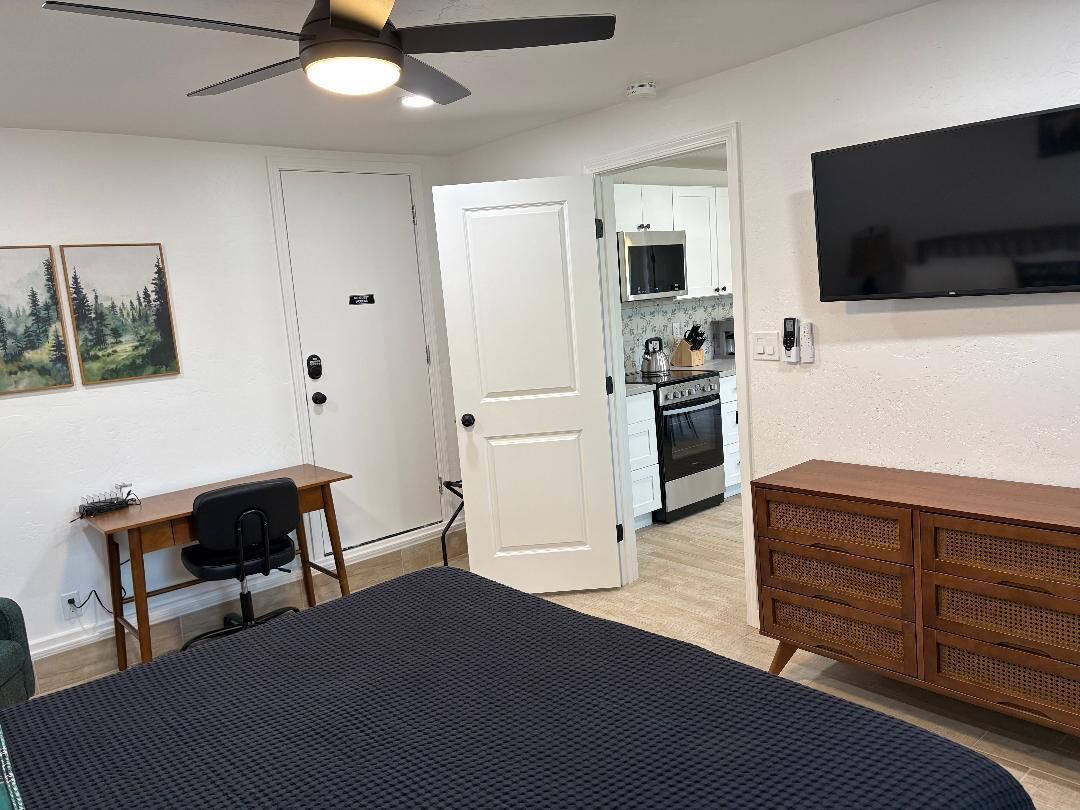 Une chambre avec un lit, un bureau, une chaise, un ventilateur de plafond, une commode et, au fond, un coin cuisine.