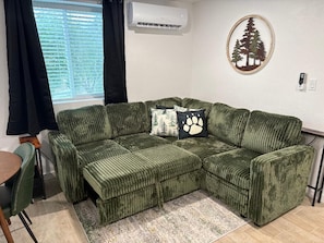 Um sofá seccional verde com manta estampada, uma mesinha, uma cadeira, um ar condicionado fixado na parede e uma silhueta decorativa de árvore.
