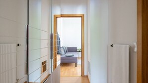 Un couloir mène à un espace de vie comprenant un canapé et une petite table, les murs sont blancs et le sol est en bois.