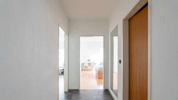 Un couloir avec une porte en bois, des murs blancs et une vue sur une pièce où se trouvent un lit et une table.