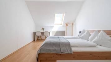 Une chambre avec un plafond en pente, un lit avec une literie blanche, un bureau avec des chaises et un radiateur.