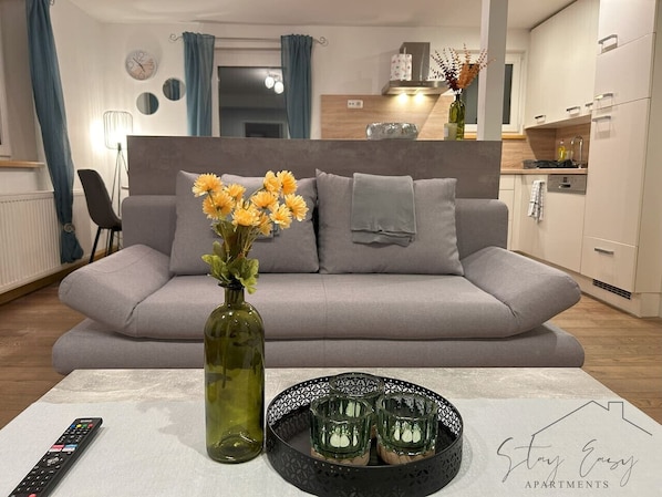 Ein Wohnzimmer mit grauem Sofa, einer Vase mit gelben Blumen, einem Tablett mit Kerzen und einer Fernbedienung auf einem weißen Tisch.