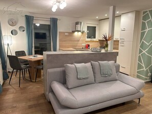 Ein modernes Wohnzimmer mit grauem Sofa, einem Esstisch mit Stühlen, einer Küchenzeile und Blick ins Freie.
