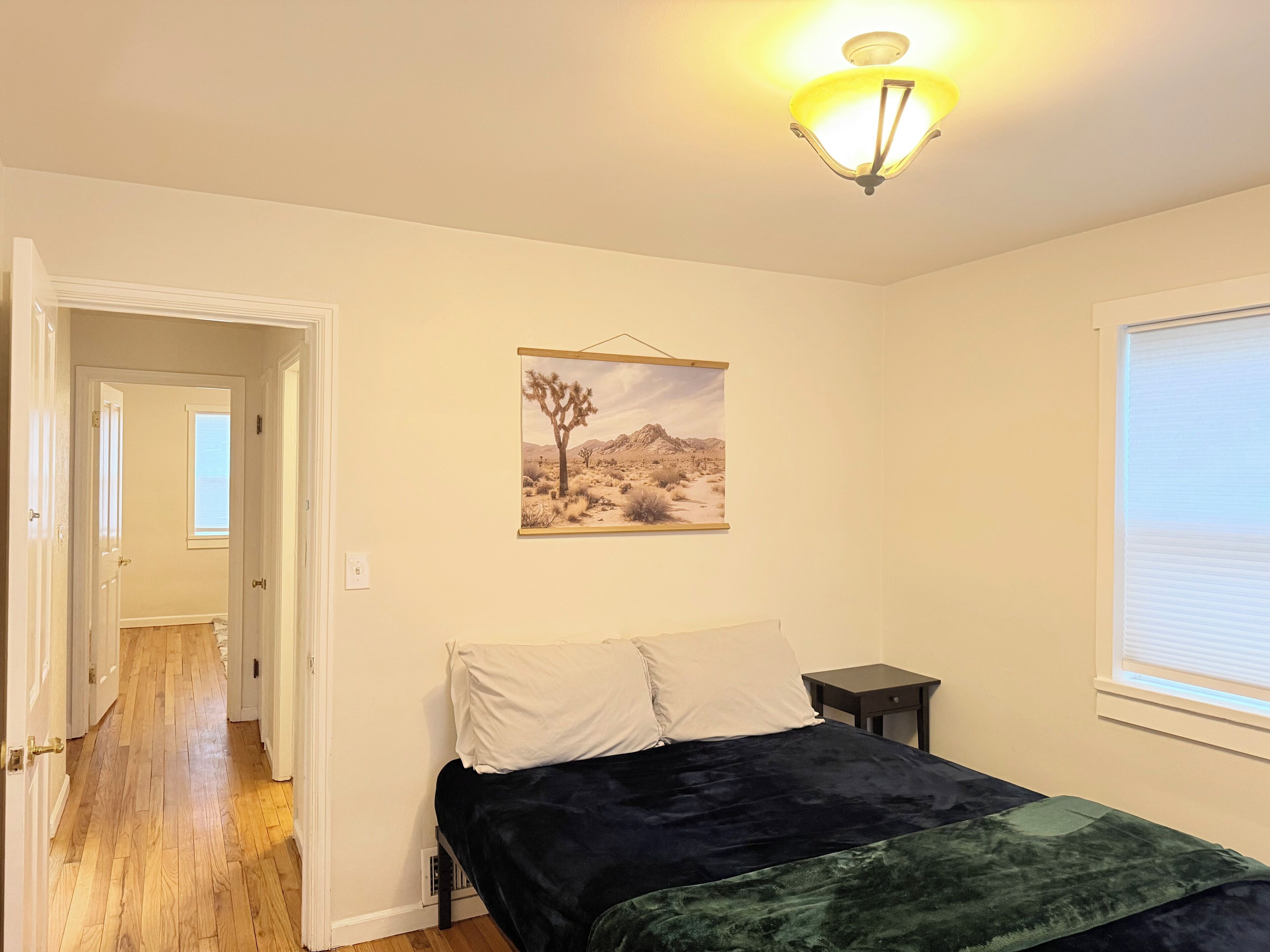 A bedroom with a bed, a nightstand, a window, and a desert landscape painting on the wall.
