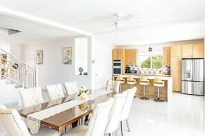Eine moderne Küche mit Edelstahlgeräten, ein Essbereich mit einem Glastisch und weißen Stühlen sowie eine Treppe mit weißer Handlauf.