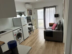 A compact living space with a kitchenette, a washing machine, a sofa, and a sliding glass door leading to a balcony.