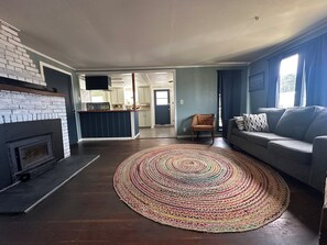 A living room with a fireplace, a sofa, a patterned rug, and a kitchen in the background.