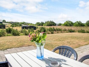 Een buiten eetgedeelte met een tafel en stoelen, een vaas met bloemen en een uitzicht op een grasveld en gebouwen op de achtergrond.