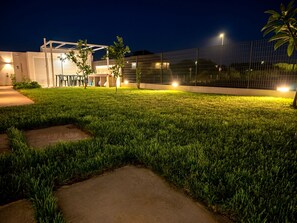 A well-lit outdoor area with a paved walkway, a small seating area, and a modern building structure. - Apartment in Villa with garden and private terrace (Kaukana)