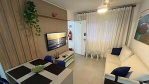 A modern living room with a sofa, a dining table with chairs, a TV mounted on the wall, and a window with curtains. - New and Cozy - near SESC (Bertioga)