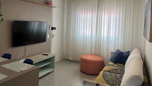 A living room with a flat-screen TV, a ceiling fan, a sofa, an ottoman, and a window with sheer curtains. - New and Cozy - near SESC (Bertioga)