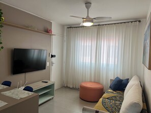 A living room with a flat-screen TV, a ceiling fan, a sofa, an ottoman, and a window with sheer curtains.
