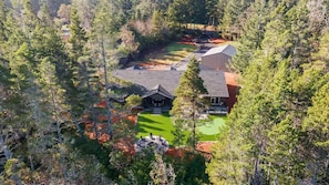 A large house surrounded by dense forest, with a well-maintained lawn and a patio area.