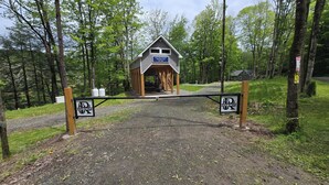 A wooden cabin with a covered porch, a gravel path leading to it, and a gate with a 'P' sign. - Rose's  RV Camping  - Private 2300' Riverfront Site on the West Branch Delaware (Starlight)