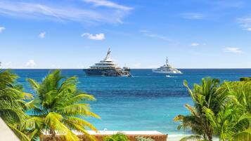 A coastal scene with two large yachts anchored in the water, surrounded by palm trees and a clear blue sky.