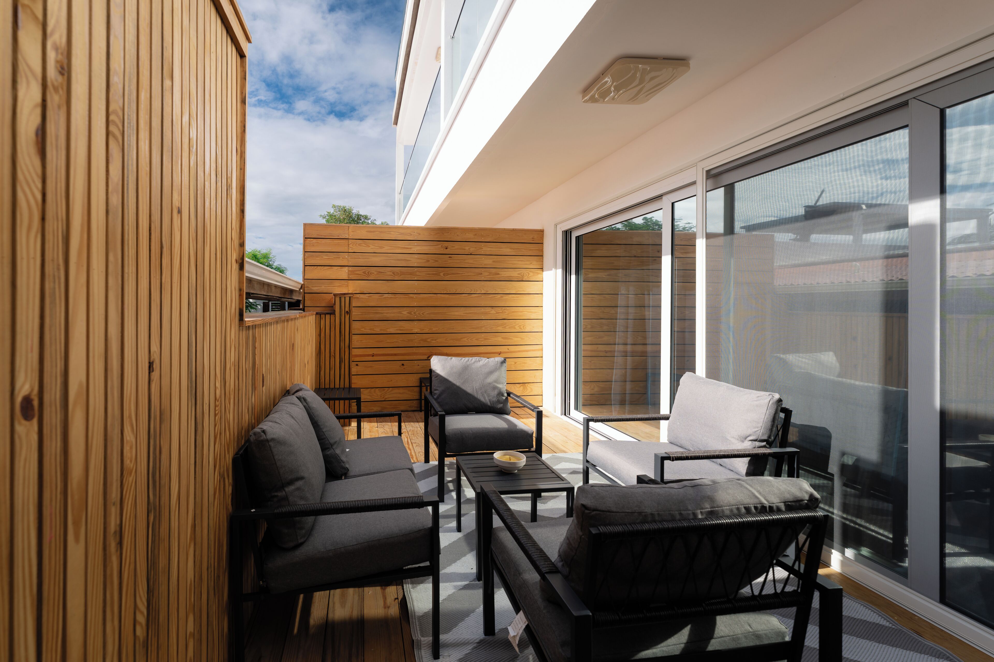 Un área de estar exterior moderna con paneles de madera, un ventanal y vista al cielo.