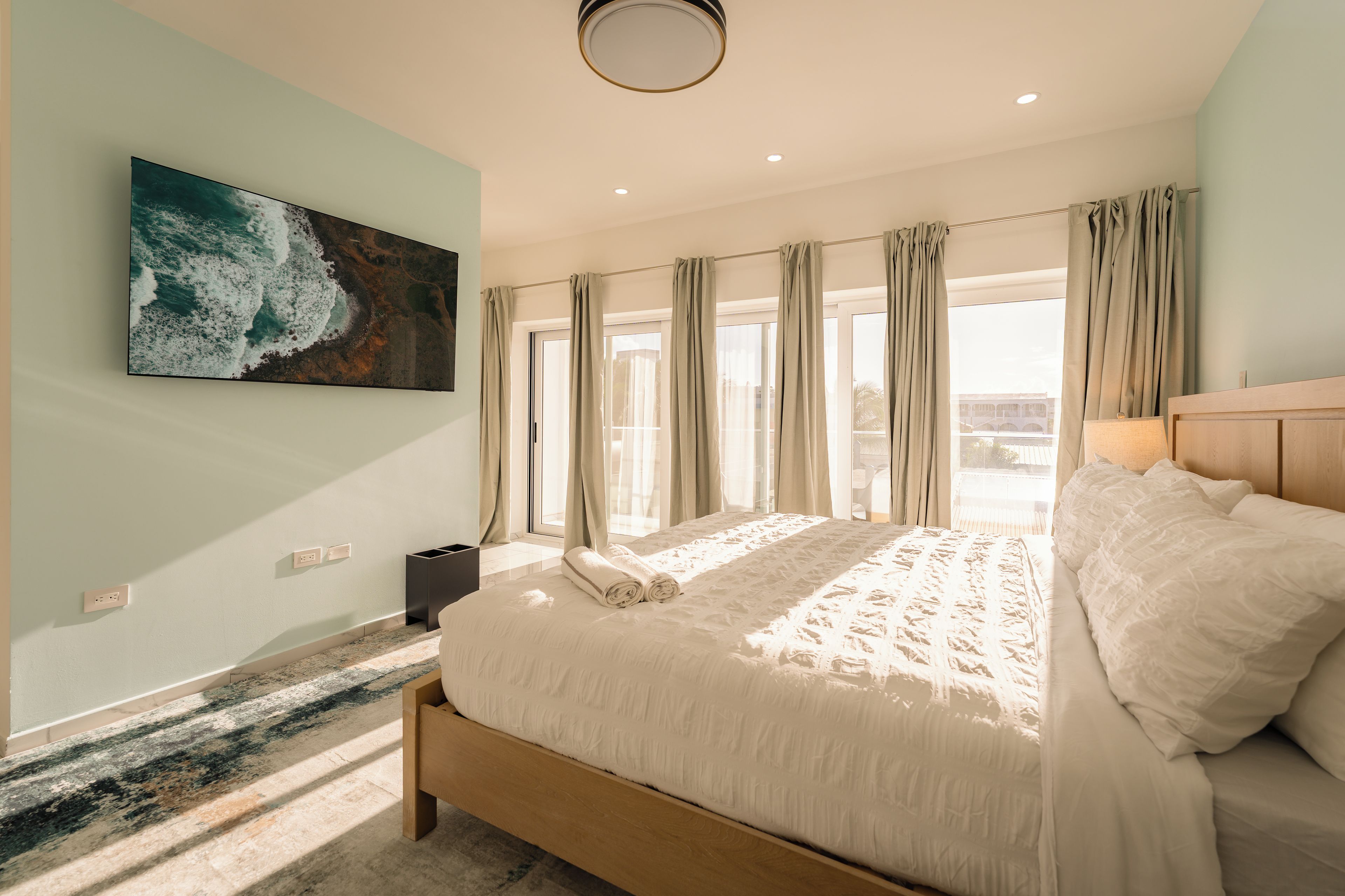 A bedroom with a large bed, a painting on the wall, and a view through the window.