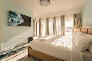 A bedroom with a large bed, a painting on the wall, and a view through the window.