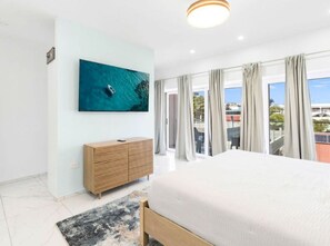 A modern bedroom with a large bed, a TV mounted on the wall, a wooden cabinet, and a view through large windows.