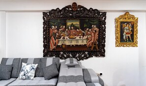 Ein Zimmer mit einem großen gerahmten Gemälde einer historischen Szene, einem Sofa mit gemusterten Kissen und einem dekorativen, goldenen Rahmen zur Rechten.
