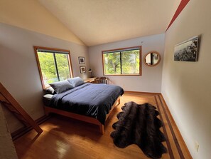 Ein Schlafzimmer mit einem Bett, zwei Fenstern, einem Schreibtisch, einem Stuhl, einem Teppich und einem Spiegel.