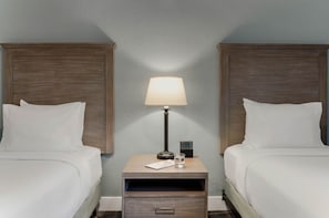 A hotel room with two beds, a bedside table with a lamp, a small drawer, and a nightstand with a clock.