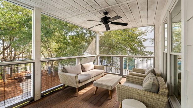 Un espacio al aire libre techado con mobiliario de mimbre, un ventilador de techo y ventanas amplias que dan a una zona boscosa.