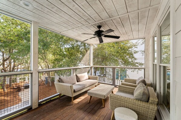 A covered outdoor area with wicker furniture, a ceiling fan, and large windows overlooking a wooded area. - Heated Pool, Lake Dock & Beach – 4BR Duplex (Inlet Beach)
