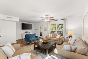 Ein Wohnzimmer mit einer Couch, einem blauen Sessel, einem Couchtisch, einem Deckenventilator, einem Fernseher und einem Fenster mit Vorhängen.